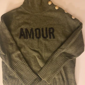 Olivgrön polotröja med text och knappar - Snygg olivgrön polotröja med svart texten 'AMOUR' framtill. Tröjan har ribbade muddar och dekorativa guldknappar längs ena axeln. Perfekt för dig som gillar trendiga detaljer och vill sticka ut lite extra. BARN STORLEK ÅR 12!!!!!