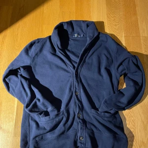 Marinblå kofta från Polo Ralph Lauren - Säljer en marinblå kofta från Polo Ralph Lauren med knäppning framtill och två fickor. Koftan har en hög krage och är tillverkad i mjuk bomull, perfekt för lager-på-lager. Klassisk och stilren modell som funkar året runt.