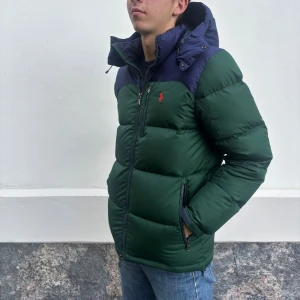 Ralph Lauren jacka  - Snygg grön pufferjacka från Polo Ralph Lauren med mörkblå axelparti och huva. Jackan har dragkedja framtill, två fickor med dragkedja och röd broderad logga på bröstet. Perfekt för kalla dagar och riktigt skön att bära.