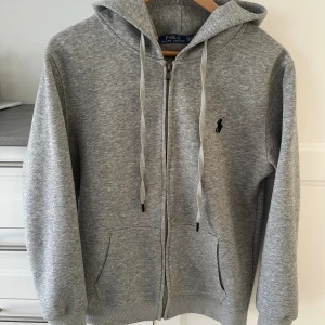 Grå hoodie från Polo Ralph Lauren - Snygg grå hoodie från Polo Ralph Lauren med dragkedja och huva. Tröjan har snörning vid halsen, ribbade muddar och fickor framtill. Klassisk svart logga broderad på bröstet. Tillverkad i mjuk bomull för skön komfort.