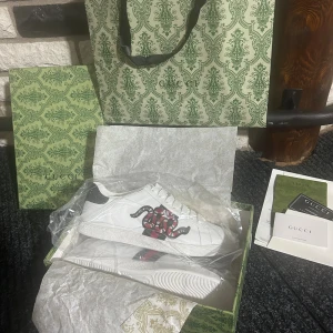 Gucci Ace sneakers - Gucci Ace sneakers i vitt skinn med klassiska gröna och röda ränder på sidan. Ena skon har en broderad röd och svart orm ovanpå ränderna. Bakkappan är i mörkgrönt, metallicmönstrat skinn med Gucci-logga. Platt sula och rund tå, snörning framtill.