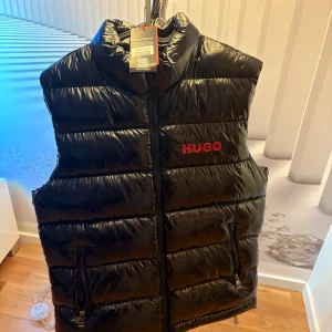 Svart dunväst från Hugo - Svart quiltad dunväst från Hugo med röd broderad logga fram och bak. Västen har hög krage, dragkedja framtill och två fickor med dragkedja. Tillverkad i glansig polyester med en clean och sportig look.