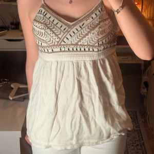 Vit broderad linnetopp från Hollister - Supersöt vit linnetopp från Hollister med broderade detaljer och pärlor på bröstet. Toppen har smala axelband, v-ringning och ett flowy fall nedtill. Har en boho-vibe med sitt mönstrade överdel. Har 2 supersmå fläckar som kan gå bort 