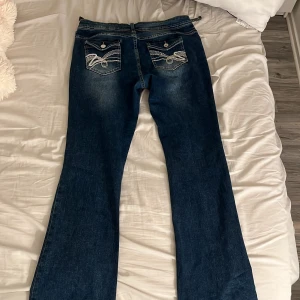 Blå bootcut jeans med broderade fickor - Snygga mörkblå bootcut jeans med coola broderade detaljer på bakfickorna och vita kontrastsömmar. Jeansen har klassisk femficksdesign, knappstängning och är tillverkade i slitstarkt denim. Perfekt för dig som gillar unik stil och bredare benslut. där man lägger bälte är trasiga på båda ställen tyvärr och en knapp är av 