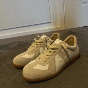 Maison Margiela Gats vit och beige  - Maison Margiela sneakers i beige och vitt med klassisk gummisula. Ovandelen är i en mix av mocka och skinn, med snörning och rund tå. Snygg minimalistisk design med Margielas ikoniska numrerade etikett på plösen. Perfekt för dig som gillar stilrena och trendiga sneakers.