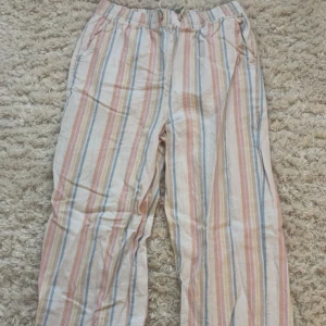 Randiga linnebyxor i pastellfärger - Säljer ett par luftiga linnebyxor med pastellfärgade ränder i rosa, blått, gult och beige. Byxorna har resår i midjan och raka ben, perfekta för varma dagar. Materialet är mjukt och svalt linne, och de har fickor på sidorna. Använda för längesedan!