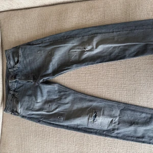 Grå slitna jeans från jack and jones - Säljer ett par grå jeans från Jack and jones med slitningar och hål på flera ställen för en cool, avslappnad look. Jeansen har klassisk femficksdesign, raka ben och är tillverkade i mjukt bomullstyg. Perfekta för dig som gillar en lite ruffare stil.