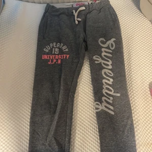 Grå mjukisbyxor från Superdry S - Mörkgrå mjukisbyxor från Superdry med vit snörning i midjan och stora tryck på benet. Byxorna har elastisk midja och är tillverkade i mjukt bomullsmaterial. Perfekta för chill eller träning, med coola Superdry-loggor och sportig vibe.