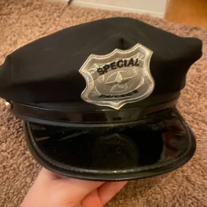 Svart polis-keps med badge - Cool svart keps med blank skärm och silvrig badge framtill där det står 'SPECIAL POLICE'. Perfekt för temafest eller maskerad. Kepsen har klassisk form och detaljer som ger en autentisk look.