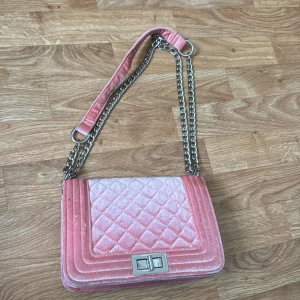 Rosa quiltad axelväska i sammet - Snygg rosa axelväska i mjuk sammet med quiltat mönster och silverfärgad kedja som axelrem. Väskan har ett stilrent lås framtill och flera fack inuti, perfekt för att hålla ordning på dina grejer. Kedjan ger en cool vibe och färgen sticker verkligen ut.