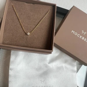 Mockberg halsband med guldton och sten - Elegant halsband från Mockberg med tunn guldfärgad kedja och en rund, glittrande sten i mitten. Perfekt för dig som gillar stilrena och klassiska smycken med en touch av lyx. Levereras i originalask.