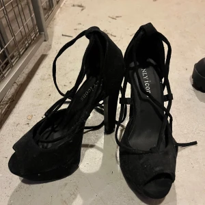 Svarta pumps med snörning från NLY ICONS - Säljer ett par svarta pumps från NLY ICONS med höga klackar och öppen tå. Skorna har snygga snörningsdetaljer som går runt vristen och är tillverkade i mockaliknande material. Perfekta för att lyfta vilken outfit som helst.