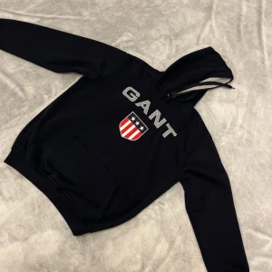 Svart hoodie från GANT med tryck - Svart hoodie från GANT med stort tryck på bröstet och ett sköldmärke i rött, vitt och blått. Tröjan har huva med snörning, magficka och ribbade muddar vid ärmslut och nederkant. Perfekt för dig som gillar klassisk streetstil. 
