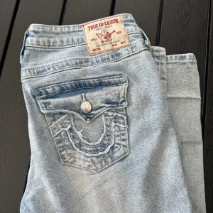 True Religion Joey Low Rise Flare Jeans - Säljer ett par ljusblå True Religion Joey jeans med låg midja och flare-ben. Klassiska detaljer med markerade sömmar och snygga fickor med lock och knapp bak. Jeansen är i bomull och har en ikonisk True Religion-patch bak i midjan.