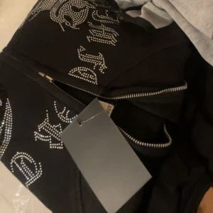 Svart hoodie med strass från aim for the moon  - Svart hoodie från aim for the moon med strassdetaljer och glittrande logotyp framtill. Tröjan har dragkedja och huva, perfekt för dig som gillar statement-plagg. Snyggt och lyxigt streetwear som sticker ut.