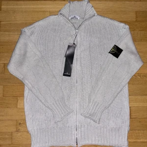 Stone Island tröja - Ljusgrå stickad kofta från Stone Island med dragkedja hela vägen och hög krage. Klassisk patch på vänster ärm och ribbade muddar. Perfekt för lager-på-lager och snygg till jeans eller cargobyxor.