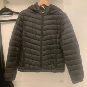 Svart pufferjacka med huva - Svart pufferjacka med huva och dragkedja framtill. Jackan har quiltad design, två fickor med dragkedja och mjukt foder på insidan. Perfekt för kyliga dagar och har en clean, stilren look.