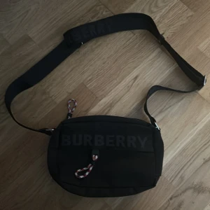 Svart axelväska från Burberry - Svart axelväska från Burberry med justerbar rem och stort Burberry-tryck på framsidan. Väskan har en rundad form, dragkedja upptill och en cool snodd-detalj i rött, vitt och svart. Perfekt storlek för dina viktigaste grejer och stilren look.