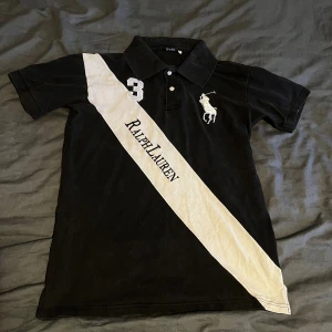 Svart Polo Ralph Lauren piké M - Svart pikétröja från Polo Ralph Lauren med vit diagonal panel och broderad logga. Snyggt nummer 3 på bröstet och klassisk krage med knappar. Materialet är 100% bomull och tröjan har korta ärmar. Stort Ralph Lauren-tryck på framsidan och detaljer på kragen. Har blivit lite solblekt men fortfarande jätte snygg. Hör av er vid frågor!