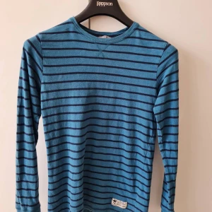 Randig långärmad tröja från Lindex strl 170 (barn) - Säljer en blå långärmad tröja med svarta horisontella ränder från Kids by Lindex. Tröjan har rund halsringning och en liten etikett nertill. 100% bomull. Storlek 170. Kan hämtas i Vallentuna.