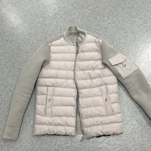 Moncler cardigan  - Snygg cardigan från Moncler med quiltad framsida och ribbstickade ärmar. Jackan har en praktisk ficka med Moncler-logga på ärmen, två sidofickor med knappstängning och dragkedja framtill. Insidan har coolt serietryck och Moncler-märke.