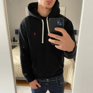 Svart hoodie från Polo Ralph Lauren - Svart hoodie från Polo Ralph Lauren med vit snörning och röd broderad logga på bröstet. Klassisk känguruficka framtill och mjukt material som känns skönt mot huden. Perfekt för dig som gillar stilren och enkel streetwear. Den är i jättebra skick, inga defekter eller skråmor. Som man ser har den dragit till sig massa hårstrån och sånt men den tvättas innan den skickas iväg! Fraktar snabbt, kom med prisförslag eller frågor.