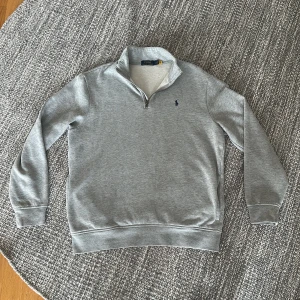 Grå half zip tröja från Polo Ralph Lauren - Snygg grå långärmad tröja från Polo Ralph Lauren med half zip och broderad marinblå logga på bröstet. Mjuk insida och klassisk passform, perfekt för lager-på-lager. Passar dig som gillar stilren och enkel design.