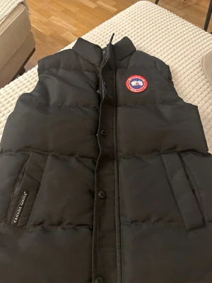 Svart dunväst från Canada Goose - Svart dunväst från Canada Goose med klassisk logga på bröstet och diskret varumärkesdetalj vid fickan. Västen har hög krage, tryckknappar och två stora fickor framtill. Perfekt för lager-på-lager under kyliga dagar.