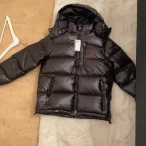 Svart dunjacka från Ralph Lauren - Svart, glansig dunjacka från Ralph Lauren med huva och dragkedja. Jackan har RL-detalj på dragkedjan och röd broderad logga på bröstet. Flera fickor med dragkedja och klassisk pufferstil. Perfekt för kalla dagar och riktigt snygg streetstil.