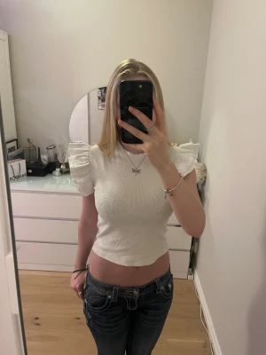 Vit croppad topp med volangärmar - Supersöt vit topp från Gina Tricot med ribbad struktur och kort, croppad passform. Toppen har rund hals och korta ärmar med volangdetaljer som ger en extra touch. Perfekt att styla med jeans eller kjol för en fräsch look. Figurnära och passar jättegulligt till sommaren. Storleken är 158/164 så den passar som en xs skulle jag säga och den är endast använd ett fåtal gånger. 