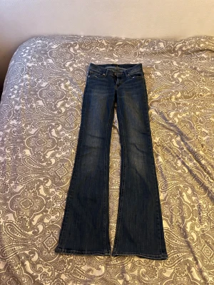 Blå bootcut jeans från Lindex, stl 34 - (Har ett hål ovanför vänster jackficka, annars i jättebra skick)       Snygga blå jeans från Lindex i bootcut-modell med klassisk femficksdesign och normal passform. Jeansen har en mörk tvätt och är tillverkade i jeansmaterial med midwaist. Perfekt för dig som gillar retrovibbar och vill ha ett par jeans med lite extra sväng på benen.