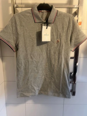 Moncler Pike - Snygg grå pikétröja från Moncler med klassisk krage och korta ärmar. Tröjan har röd och vit kant på krage och ärmslut samt Moncler-logga på bröstet. Tillverkad i mjuk bomull som känns skön mot huden. Perfekt för dig som gillar stilrena och sportiga plagg.