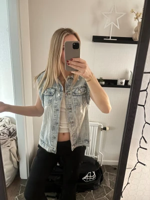 Ljusblå jeansväst  - Säljer en snygg ljusblå jeansväst med klassisk krage och silverfärgade knappar framtill. Västen har två bröstfickor med lock och är ärmlös, perfekt att slänga över en t-shirt eller hoodie för en chill look. Passar dig som gillar street och denim. Västen är från shine men otroligt bra skick använd ca 5-10 ggr, nypris:150kr 