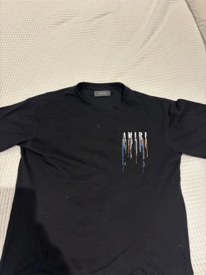 Svart AMIRI t-shirt med färgstänk - Svart t-shirt från AMIRI med tryckt logga och färgstänk i vitt, blått, orange och beige både fram och bak. Klassisk rund hals och korta ärmar. Skön passform och tillverkad i mjuk bomull. Perfekt för dig som gillar streetwear med en konstnärlig twist.