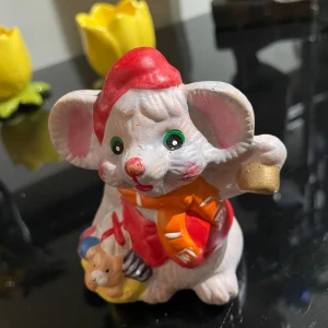 Söt musfigur elefant i keramik med röd mössa - Charmig liten musfigur elegant i keramik, ca 10 cm hög. Musen har grönskimrande ögon, röd mössa och röd väst, samt en orange halsduk med vita detaljer. Den håller en guldfärgad klocka och har en nalle bredvid sig. Perfekt som dekoration eller present.