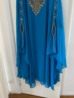 Blå långklänning med paljetter - Säljer en blå långklänning i flowigt tyg med extravagant paljettdekor i guld och silver över bröstet och längs ärmarna. Klänningen har ett unikt, svepande fall och breda ärmar som ger en riktigt lyxig vibe. Perfekt för dig som vill sticka ut med glittriga detaljer.