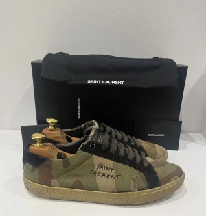 Saint Laurent camo sneakers - Sällsynta sneakers från Saint Laurent med coolt camouflage-mönster i grönt, beige och brunt. Skorna har svarta detaljer, snörning och rund tå. Materialet är canvas med mockadetaljer och Saint Laurent-logga på sidan. Perfekt för dig som gillar streetstyle och designer.
