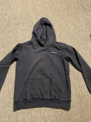 Mörkblå hoodie Peak Performance - Svart hoodie från Peak Performance i storlek 160. Klassisk modell med huva, magficka och diskret logga på bröstet. Mjuk insida och ribbade muddar vid ärmslut och nederkant. Perfekt för chill eller skola. Storlek 160.