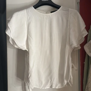 Vit blus med volangärm och knyt - Superfin vit blus från Zara med volangärmar och knytband i sidorna. Blusen har rund halsringning och är tillverkad i ett lätt, lite glansigt material som ger en lyxig känsla. Perfekt för dig som gillar romantiska detaljer och en luftig look. Sydd på sidorna på tröjan men syns inte 