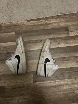 Nike Blazer Mid vit/svart skinn - Nike Blazer Mid sneakers i vitt skinn med svart swoosh på sidan. Klassisk high-top modell med snörning och perforerade detaljer på tån. Platt sula och rund tå. Snygg retrostil som passar till streetwear och casual outfits.