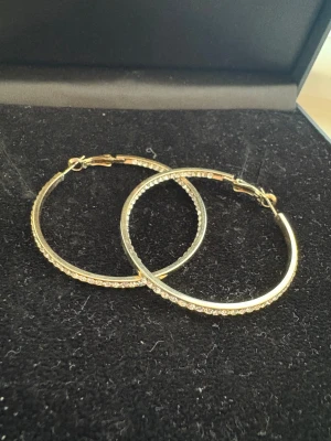 Glittriga stora guldfärgade hoops - Säljer ett par stora, runda örhängen i guldfärg med glittrande stenar längs hela bågen. De har klassisk hoop-form och stängs med ett smidigt lås. Perfekta för dig som vill ha lite extra bling och synas i mängden.