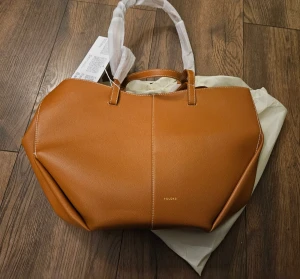 Brun handväska från Polène i skinn - Snygg handväska från Polène i brun färg med stilren, geometrisk form. Väskan är tillverkad i skinn med vita kontrastsömmar och har både korta och långa handtag. Perfekt för dig som gillar minimalistisk design och vill ha en rymlig väska med exklusiv känsla.