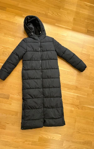 Svart lång pufferjacka med huva - Säljer en svart lång pufferjacka från Cubus med huva och dragkedja hela vägen. Jackan är quiltad och har en clean look med raka linjer och långa ärmar. Perfekt för kalla dagar när du vill hålla dig varm och snygg. Köpt för 850, säljer för 600. (Använd två gånger bara)