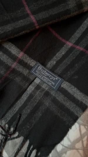 Rutig svart halsduk från Burberrys - Snygg halsduk från Burberrys i klassiskt rutigt mönster med svarta, gråa och röda toner. Gjord i 100% lammull och har fransar i kanterna. Perfekt accessoar för att lyfta din vinterstil med en tidlös vibe. 