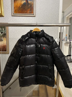 Svart Polo Ralph Lauren jacka M - Säljer en svart pufferjacka från Polo Ralph Lauren med röd logga på bröstet. Jackan har huva, dragkedja framtill och två sidofickor med dragkedja. Den är quiltad och har en glansig finish som ger en cool vibe. Perfekt för kalla dagar och riktigt snygg till streetwear.