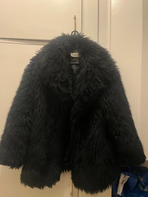 Svart pälsjacka från OuiSet - Säljer en svart fluffig pälsjacka från OuiSet i storlek 38. Jackan har en oversized look med långärmade ärmar och är tillverkad av mjuk modakryl. Perfekt för dig som vill ha en statement piece med mycket attityd och volym. Innerfodret i ena armen behövs sys, se bilder i annonsen.