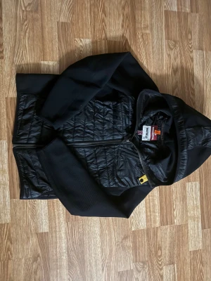 Svart quiltad jacka Parajumpers Y-M - Svart jacka från Parajumpers med huva, quiltad framsida och detaljer i orange och gult. Jackan har dragkedja, broderad patch på ärmen och är tillverkad i syntetmaterial med stickade partier på ärmarna. Perfekt för dig som gillar street och sportig stil.