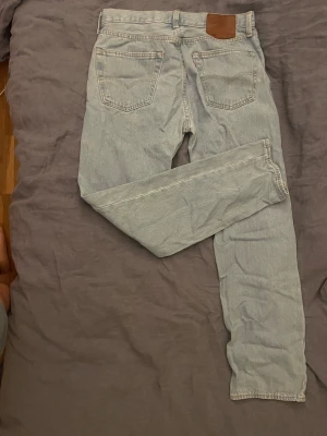 Levi's 501 ljusblå jeans - Ljusblå Levi's 501 jeans med klassisk rak passform och fem fickor. Jeansen har en brun läderpatch bak i midjan och knappgylf framtill. Tillverkade i slitstarkt bomullsmaterial, perfekt för dig som gillar tidlös denimstil.