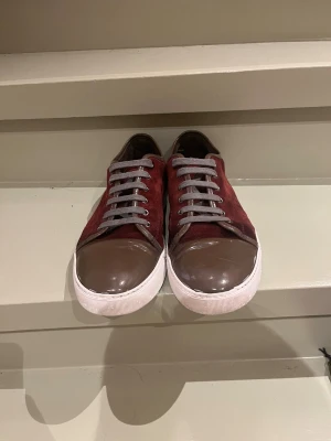 Vinröda sneakers med bruna detaljer - Snygga vinröda sneakers med ovandel i mocka och grön lackade tår. Skorna har grå snörning, vita sulor och bruna detaljer runt kanterna. Perfekta för dig som vill ha en unik och stilren look till vardags. Storleken är UK 10. Självklart så ingår box samt dustbags. Nypris: 5000kr. Vårat pris: 1699kr. 