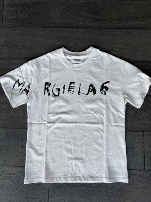 Vit Margiela t shirt  - Säljer en vit t-shirt från MM6 Maison Margiela med svart, handskrivet tryck över bröstet och ärmen. T-shirten har rund hals, rak passform och är tillverkad i mjuk bomull. Perfekt statement-plagg för dig som gillar unik design.